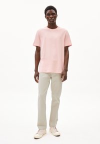 Lichtroze katoenen t-shirt met korte mouwen, gecombineerd met lichtgrijze broeken en beige sneakers, staande tegen een witte achtergrond.
