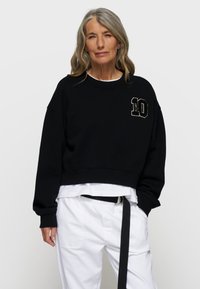 Czarny skrócony sweatshirt z okrągłym dekoltem i haftowanym numerem "10" na piersi. Nosi się go na białej warstwowej koszuli i białych spodniach.