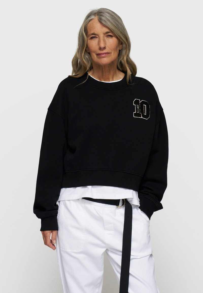 Czarny skrócony sweatshirt z okrągłym dekoltem i haftowanym numerem "10" na piersi. Nosi się go na białej warstwowej koszuli i białych spodniach.