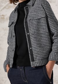 Gray quilted jacket with a button-up front, two chest pockets, black accents, and a smooth texture, paired with a black top and denim jeans.