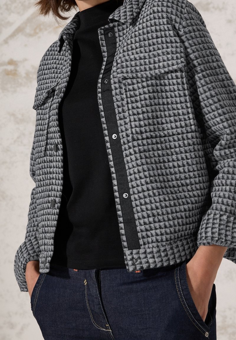 Gray quilted jacket with a button-up front, two chest pockets, black accents, and a smooth texture, paired with a black top and denim jeans.