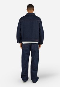 Donkerblauwe denimjack met een losse pasvorm, voorzien van knoopsluitingen en zakken. Gepaard met bijpassende losse cargobroek, ook in donkerdenim.