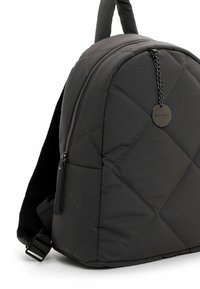 Schwarzer gesteppter Rucksack mit vorderer Reißverschlusstasche, gepolsterten Trägern und rundem Metallanhänger, der an einer Kette nahe der Oberseite hängt.