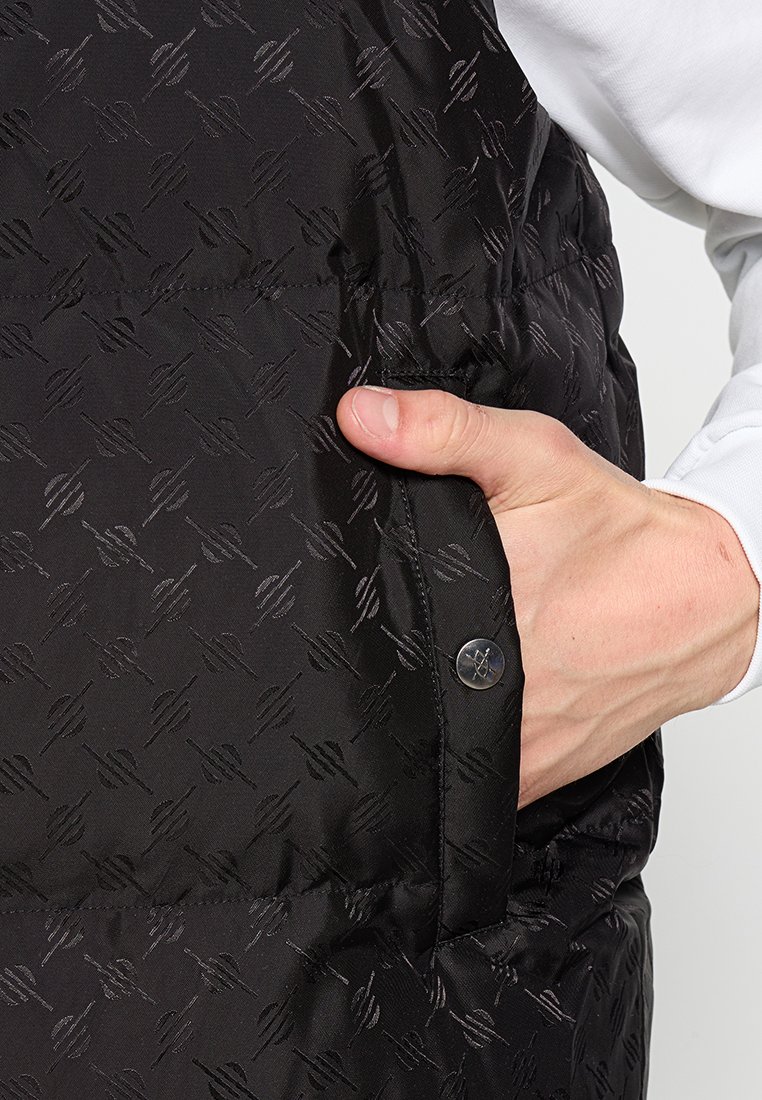 Zwarte puffer vest met een textuurpatroon, zijzak en details in zilver met drukknopen. De stof heeft een gladde, glanzende afwerking.