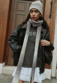 Brown leather jacket with a grey scarf and hat, layered over a dark shirt and white skirt. Features a textured, ribbed beanie and scarf.