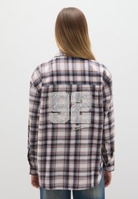 Camicia flanella a scacchi nei toni del rosa, blu e marrone presenta una grande patch con il numero "92" e la scritta "New York" sul retro. Maniche lunghe e polsini con bottoni.