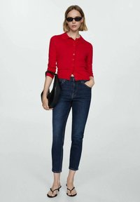 Cardigan lungo rosso con maniche lunghe e bottoni, indossato con jeans a vita alta blu scuro e sandali neri con tacco, completato da occhiali da sole scuri.