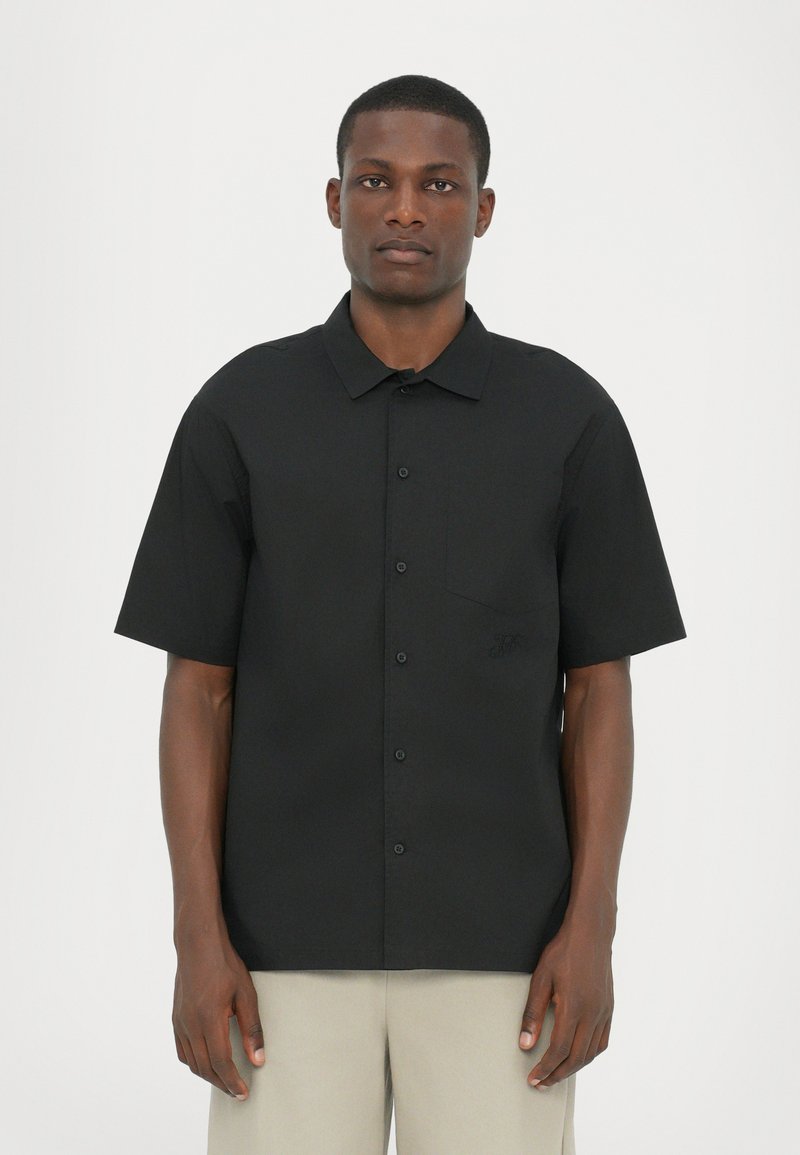 Black short-sleeve shirt in a plain design with a chest pocket and subtle logo detail. Fabric appears smooth, with a relaxed fit.