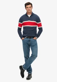 Suéter de punto navy con rayas horizontales rojas y blancas, cuello con cremallera, combinado con jeans azules y botas negras hasta el tobillo.
