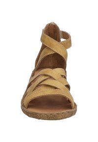 Keltainen nahkainen sandaali, jossa on ristikkäissuunnittelu, avonainen varvas ja teksturoitu pohja. Beigen väriset ompeleet korostavat hihnoja. Sopii lämpimille säille.