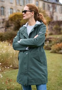 Vrouw met rood haar in een groene jas met capuchon en zonnebril staat met gekruiste armen in een tuin met een historisch stenen gebouw op de achtergrond.