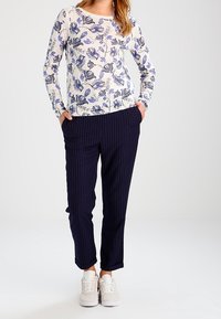 Frau trägt ein weißes, blumenmusterte Langarmoberteil, marineblaue gestreifte Hose mit hochgekrempelten Säumen und weiße Sneakers, steht mit den Händen in den Taschen.