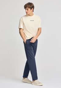 Camiseta beige con "POLO CLUB" impreso en negro, combinada con pantalones de marinero oscuros y zapatillas de color claro, con un ajuste relajado.