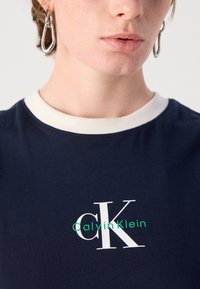 Námornícky tričko s bielym okrúhlym výstrihom, na prednej strane s bielou a zelenou grafickou potlačou "ck" a "CalvinKlein". Mäkká textúra tkaniny.