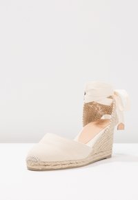 Beige kanvas espadrillo kile med en teksturert vevd hæl og bånd rundt ankelen. Har en rund tå og en jute-bekledd plattform.