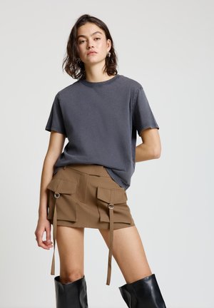 T-shirt grigia a maniche corte abbinata a una minigonna marrone con tasche frontali, dettagli in metallo a forma di anello e lacci regolabili. Stivali neri al ginocchio.