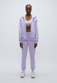 Súprava mikiny a teplákov v fialovej farbe z mäkkej látky, s predným zipsom, kapucňou a elastickými manžetami, doplnená o biele tenisky.