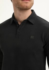 Zwart langarm poloshirt met een gestructureerde stof, knoopsluiting en een klein logo patch op de borst.