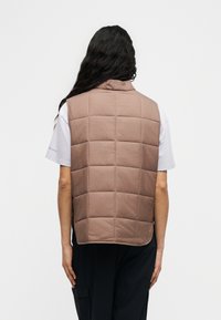 Άτομο με μακριά σκούρα μαλλιά που φοράει ανοιχτό καφέ καπιτονέ γιλέκο πάνω από λευκό κοντομάνικο πουκάμισο και μαύρο παντελόνι, με την πλάτη γυρισμένη σε λευκό φόντο.