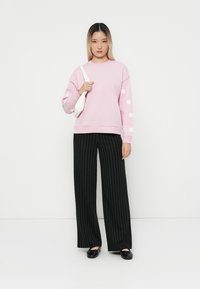 Rosa sweatshirt med blommönster på ärmarna, svarta vertikalt randiga vida byxor och svarta platta skor. Vit handväska.