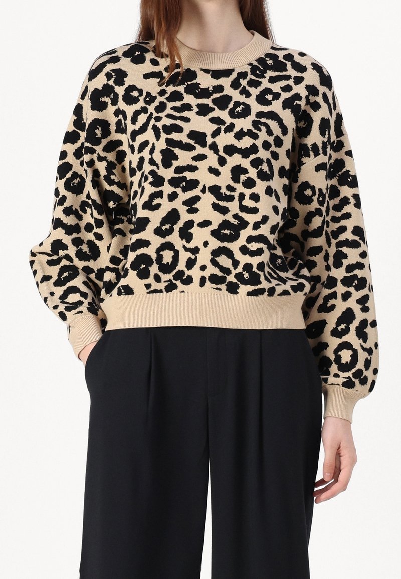 Femme portant un pull beige et noir à imprimé léopard avec manches longues et pantalon noir à jambes larges, mains dans les poches.