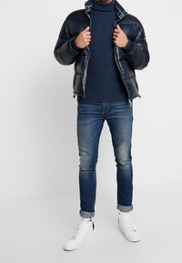 Pufferjacka i marinblått, stickad mörk tröja, jeans och vita sneakers. Har blixtlås och uppvikta jeansfållar.