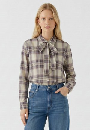 Frau mit blondem Haar, die eine beige-lila karierte Bluse mit einer Halschleife und blaue hochtaillierte Jeans trägt, steht vor einem schlichten Hintergrund.