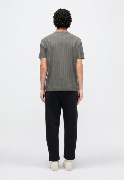 Γκρι υφασμάτινο κοντομάνικο πουκάμισο με στρογγυλή λαιμόκοψη, συνδυασμένο με μαύρο παντελόνι σε χαλαρή γραμμή. Το μοντέλο εμφανίζεται από πίσω. Ορατά λευκά αθλητικά παπούτσια.