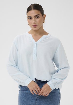 Kobieta w złotych kołach kolczyków, ubrana w jasnoniebieską, zapinaną na guziki bluzkę i niebieskie dżinsy, stojąca na jednolitym tle.