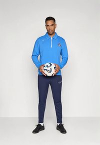 Hombre con camiseta deportiva azul y pantalones marinos, sosteniendo un balón de fútbol Nike blanco y azul, de pie frente a un fondo claro y liso.