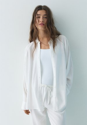 Ung kvinde med langt, løst hår iført løs hvid skjorte med knapper, hvid tanktop og hvide bukser med snor foran mod en lys baggrund.