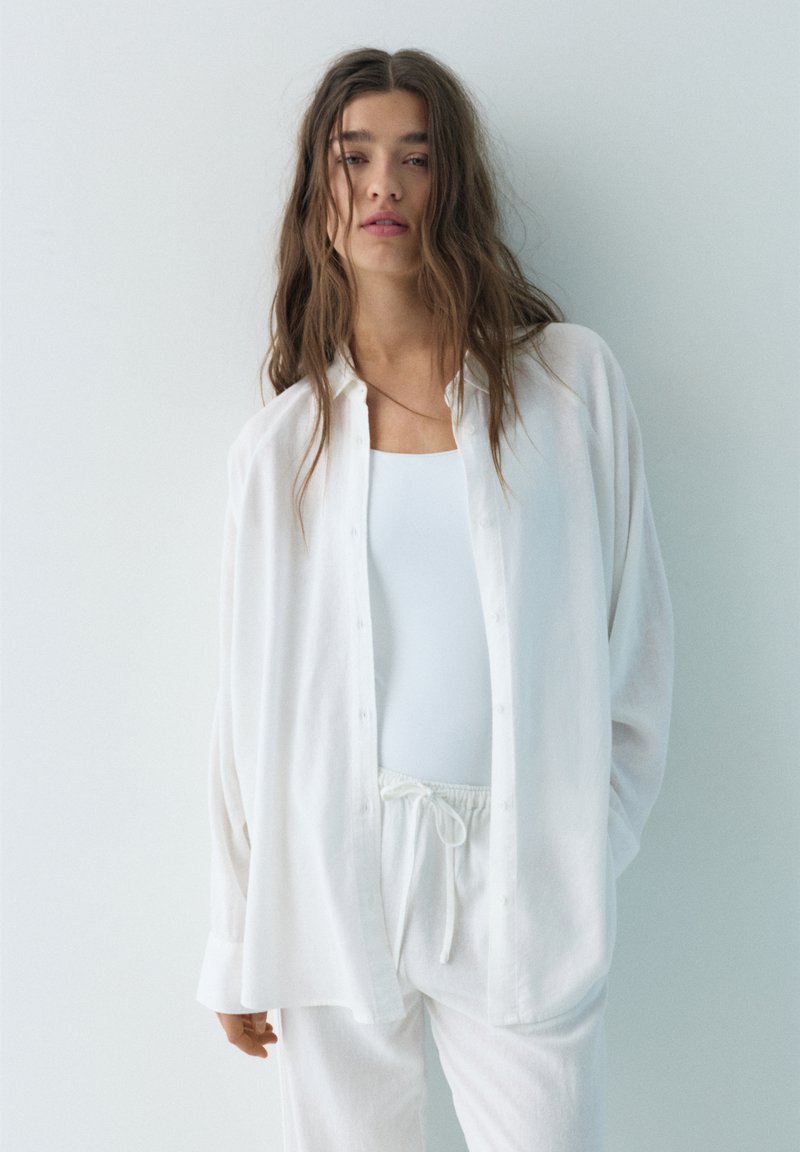 Jeune femme aux longs cheveux lâchés portant une chemise blanche ample à boutons, un débardeur blanc et un pantalon blanc à cordon de serrage sur un fond clair.