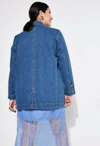 Chaqueta de mezclilla en un azul medio, con un corte recto, mangas largas y detalles de botones en los puños. Llevada sobre un vestido de encaje azul claro.
