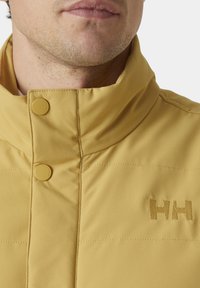 Chaqueta acolchada amarilla con cuello alto, dos botones a presión y logo bordado "HH" en el lado izquierdo del pecho. Textura suave y material ligero.