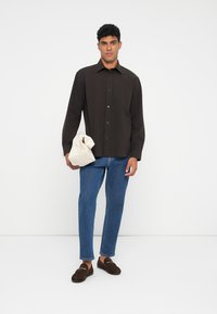 Mørkebrun skjorte med knapper og krage, kombinert med blå denimjeans. Holder en kremfarget veske, og har på seg brune loafers.
