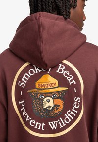 Felpa cappuccio color bordeaux con una grande grafica circolare di Smokey Bear con un cappello giallo. Il testo recita "Smokey Bear - Prevenire gli incendi boschivi."