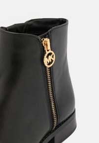 Bottine en cuir noir avec fermeture éclair dorée et tirette circulaire avec logo MK sur fond blanc.