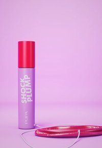 Fioletowy cylindryczny pojemnik z czerwonym korkiem oznaczony napisem "SHOCK PLUMP", obok błyszczącego różowego okrągłego przedmiotu z białym przewodem.