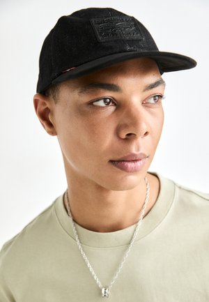Svart keps med en texturerad yta och ett framtida märke. I kombination med en ljusgrön crewneck t-shirt och ett silverfärgat kedjehalsband.