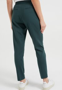 Person in dunkelgrünen maßgeschneiderten Hosen und weißen Sneakern, stehend vor einem schlichten weißen Hintergrund, von hinten gezeigt.