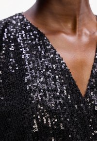 Gros plan sur une personne portant un haut noir à col en V couvert de sequins noirs brillants sur un tissu texturé.