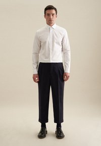 Witte overhemd met een spreadkraag, gecombineerd met donkerblauwe geplooide broek en zwarte leren schoenen, op een neutrale achtergrond.