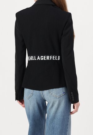 Blazer noir à rayures fines avec une silhouette ajustée, featuring une bande blanche de marque autour de la taille et des accents de boutons sur les manches. Jean bleu visible.