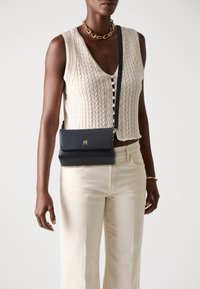 Navy leather crossbody bag with a gold logo, rectangular shape, and flap closure. Worn over a cream textured knit top and light pants.