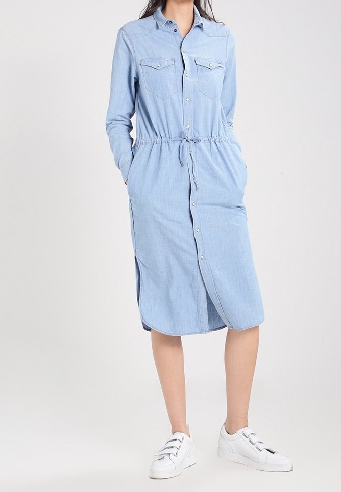 Femme portant une robe midi bleu clair à manches longues avec boutons, poches, taille avec cordon, et des baskets blanches.