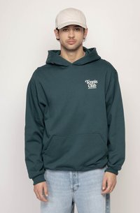 Tyrkysová mikina s kapucňou a predným vreckom, s bielym logom "Tennis Club". Dlhé rukávy s rebrovanými manžetami. Nosená s svetlomodrými džínami a béžovým klobúkom.