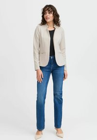 Beige kavaj med en figursydd design, två framfickor och subtil matt textur, kombinerad med mörkblå rakbenade jeans och beiga klackar.