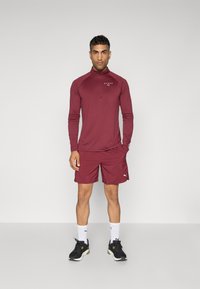 Bordowy top sportowy z półgolfem, długimi rękawami, pasujące bordowe szorty, czarne sneakersy i białe skarpetki z logo, na szarym tle.