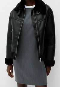 Zwarte leren jas met een bontkraag en -manchetten; zilveren ritssluiting; gedragen over een grijze jurk met een gladde textuur en een aansluitend ontwerp.