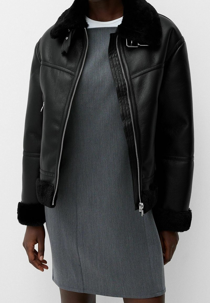 Zwarte leren jas met een bontkraag en -manchetten; zilveren ritssluiting; gedragen over een grijze jurk met een gladde textuur en een aansluitend ontwerp.